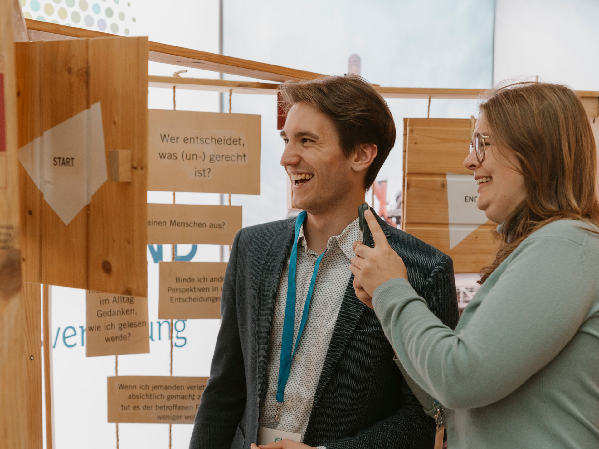27 - 25_11_Wick_Youpan_BNEWinterfest_Edits_27 Zwei junge Menschen schauen sich eine interaktive Ausstellung an und grinsen.