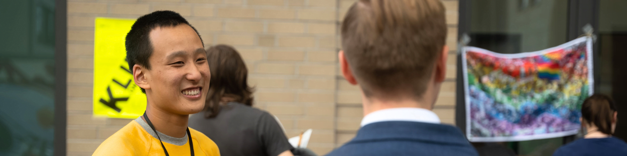 Ein junger Mensch in einem gelben T-Shirt grinst eine andere Person an, die gegenüber steht. Hinter den beiden hängen Poster und Banner, u.a. mit der Regenbogenflagge.