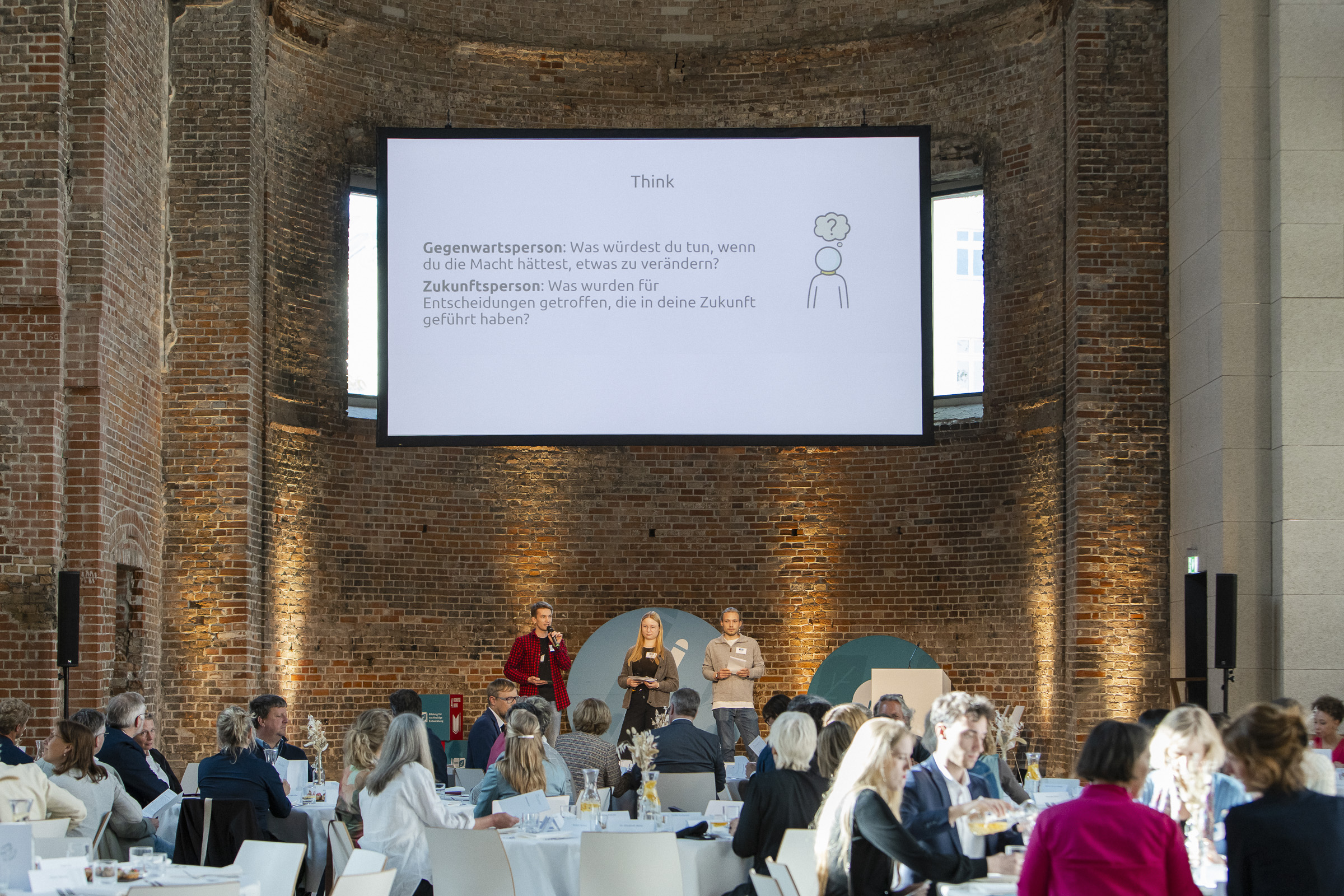 Viele Menschen sitzen in einer Kirche an runden Tischen. Im Fokus sind drei junge Menschen auf der Bühne, über ihnen wird eine Präsentation übertragen. Text: "Think. Gegenwartsperson: Was würdest du tun, wenn du die Macht hättest, etwas zu verändern? Zukunftsperson: Was wurden für Entscheidungen getroffen, die in deine Zukunft geführt haben?"
