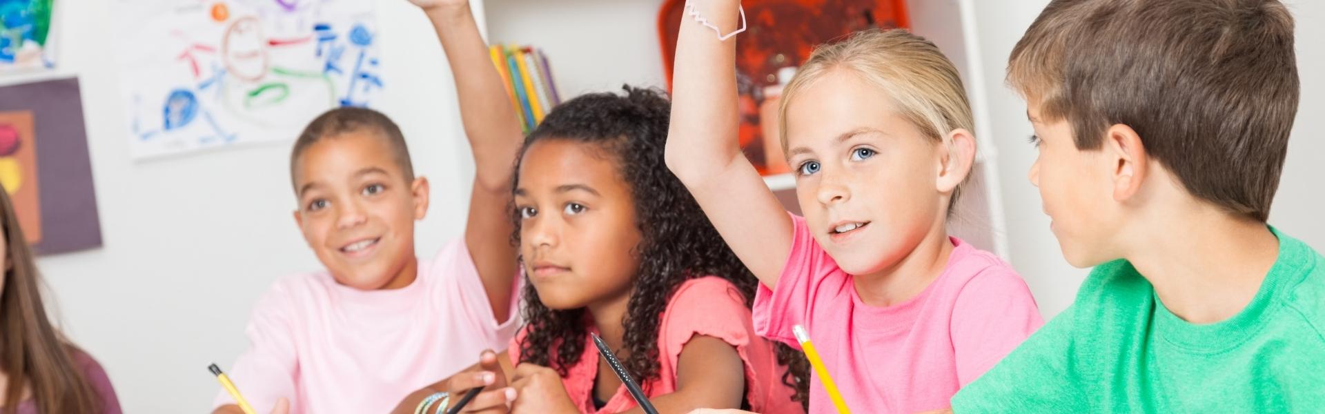 Vier Kinder sitzen nebeneinander in der Schule und zwei heben ihre Hände