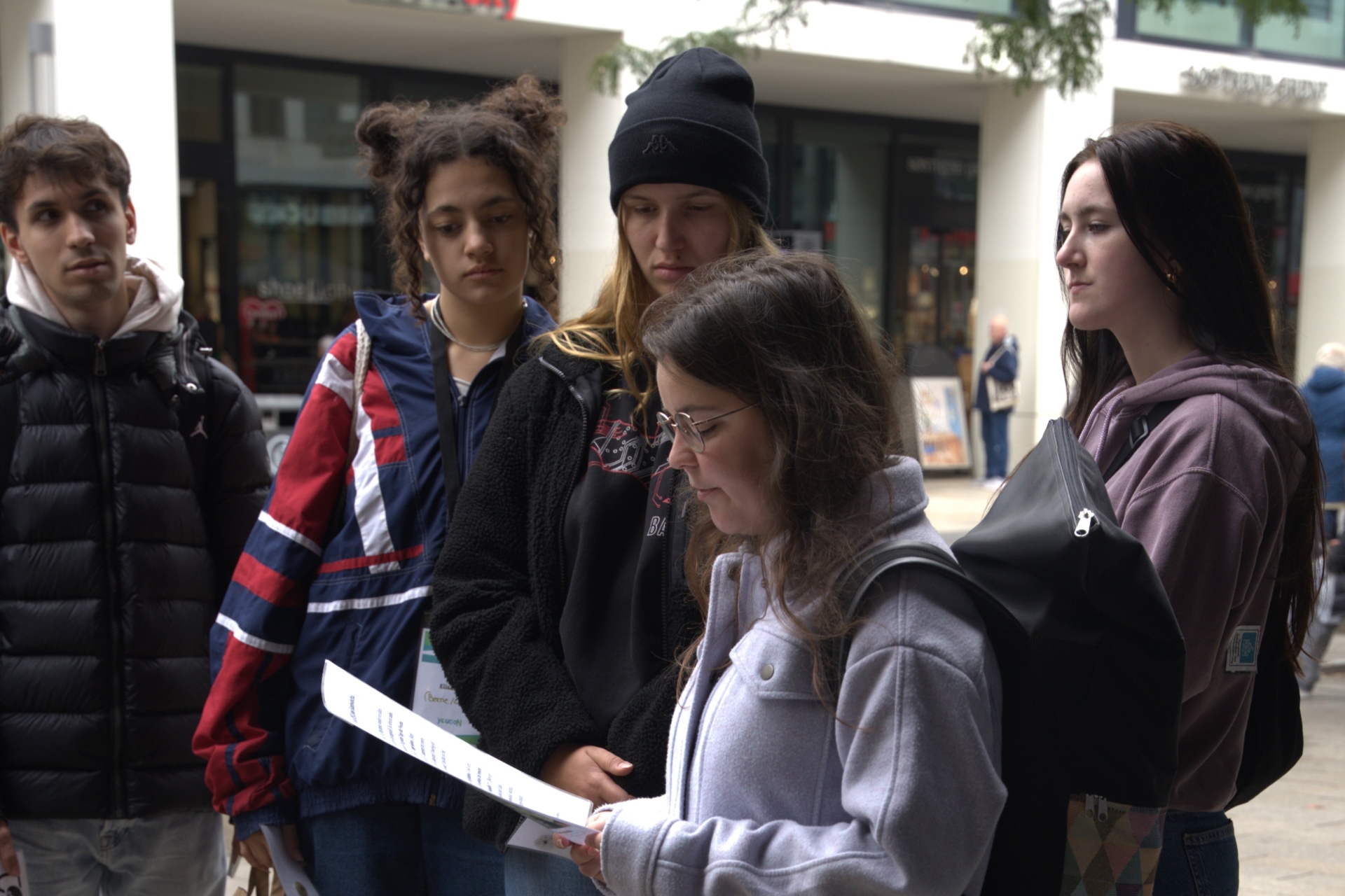 Eine junge Person steht in einer Fußgängerzone und liest ein Blatt Papier. Um sie herum stehen vier andere Junge Menschen.