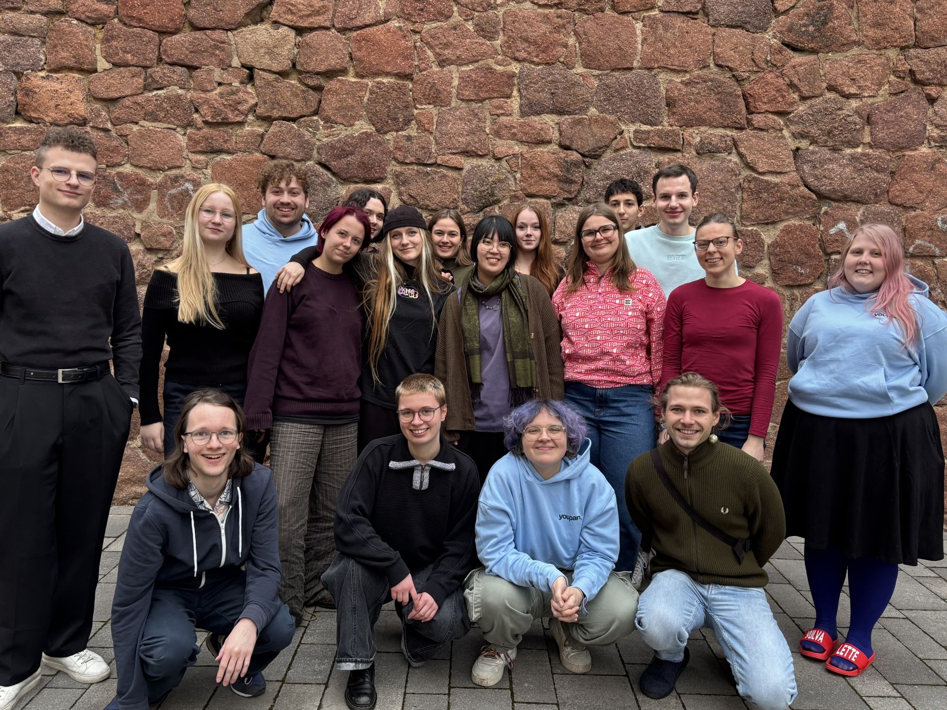 18 junge Menschen, die Mitglieder des youpan, stehen in zwei Reihen vor einer Steinmauer für ein Gruppenfoto.