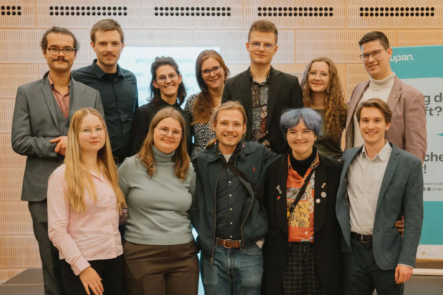 Zwölf Mitglieder des youpan stehen in einem Veranstaltungssaal auf einer Bühne aufgereiht für ein Gruppenbild.
