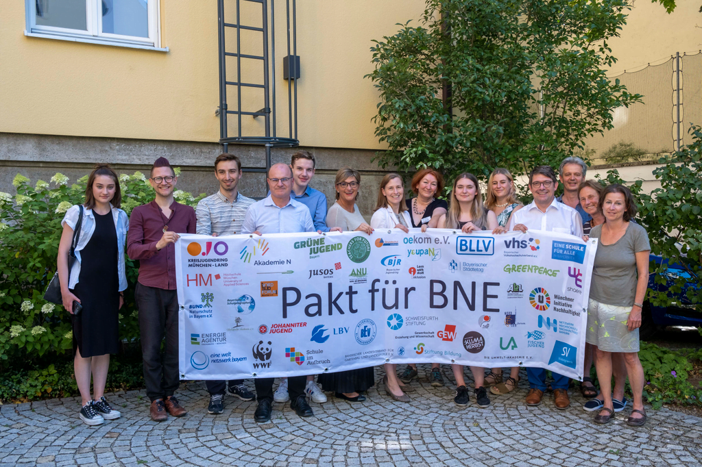Viele Menschen halten ein Banner mit bunten Logos der Unterzeichner*innen des Pakts für Bildung für nachhaltige Entwicklung in Bayern darauf hoch.