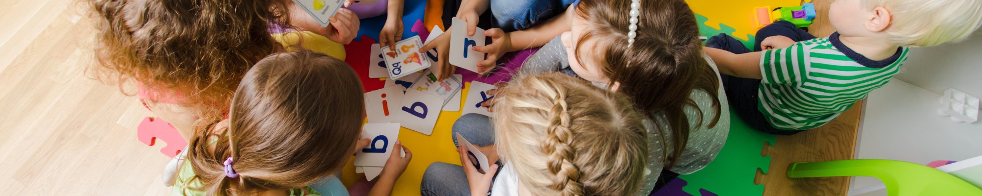mehrere Kindergarten-Kinder sitzen im Kreis auf dem Fußboden und halten Karten mit einzelnen Buchstaben in Händen